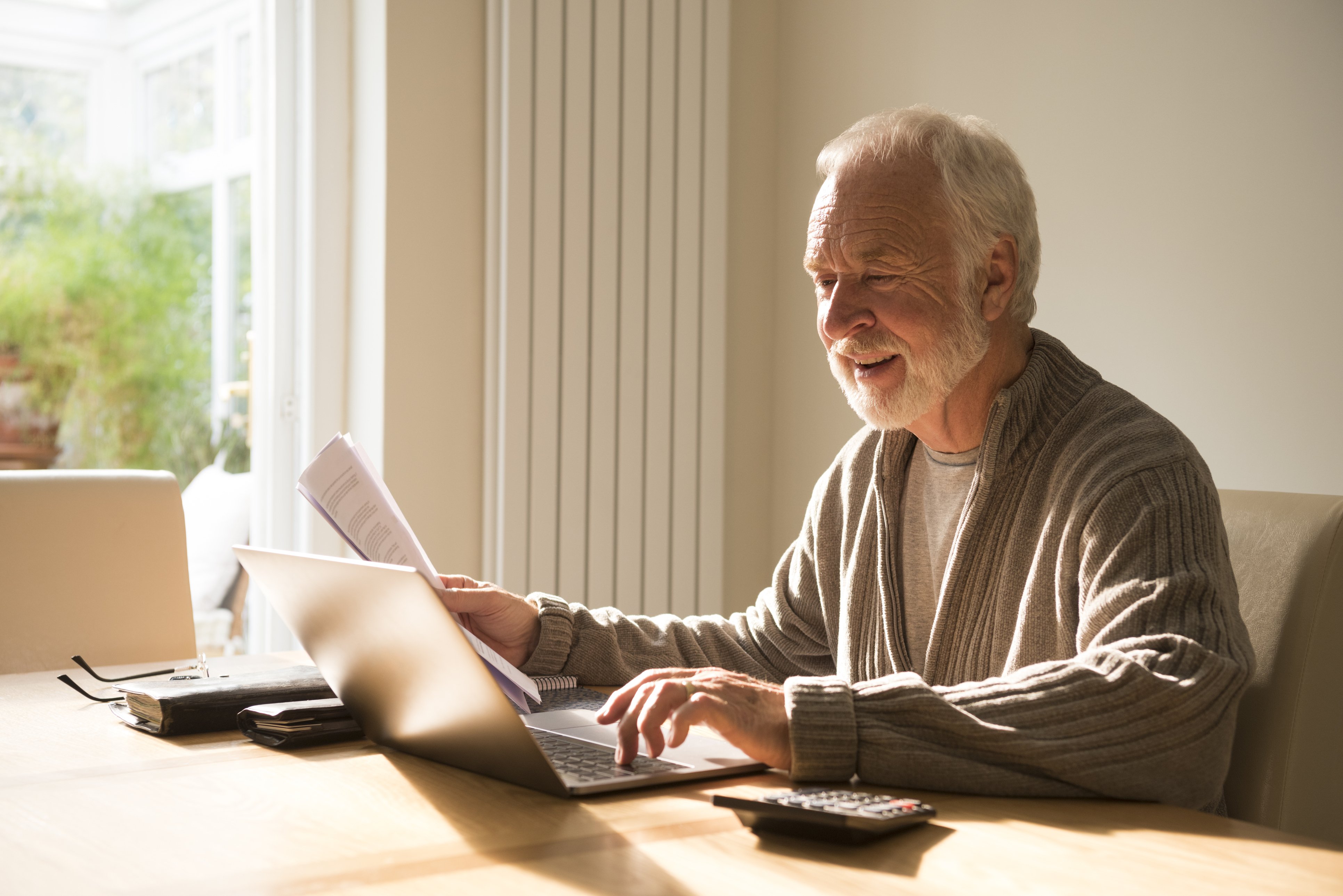 An older man looking at a laptop whilst also holding papers in his right hand