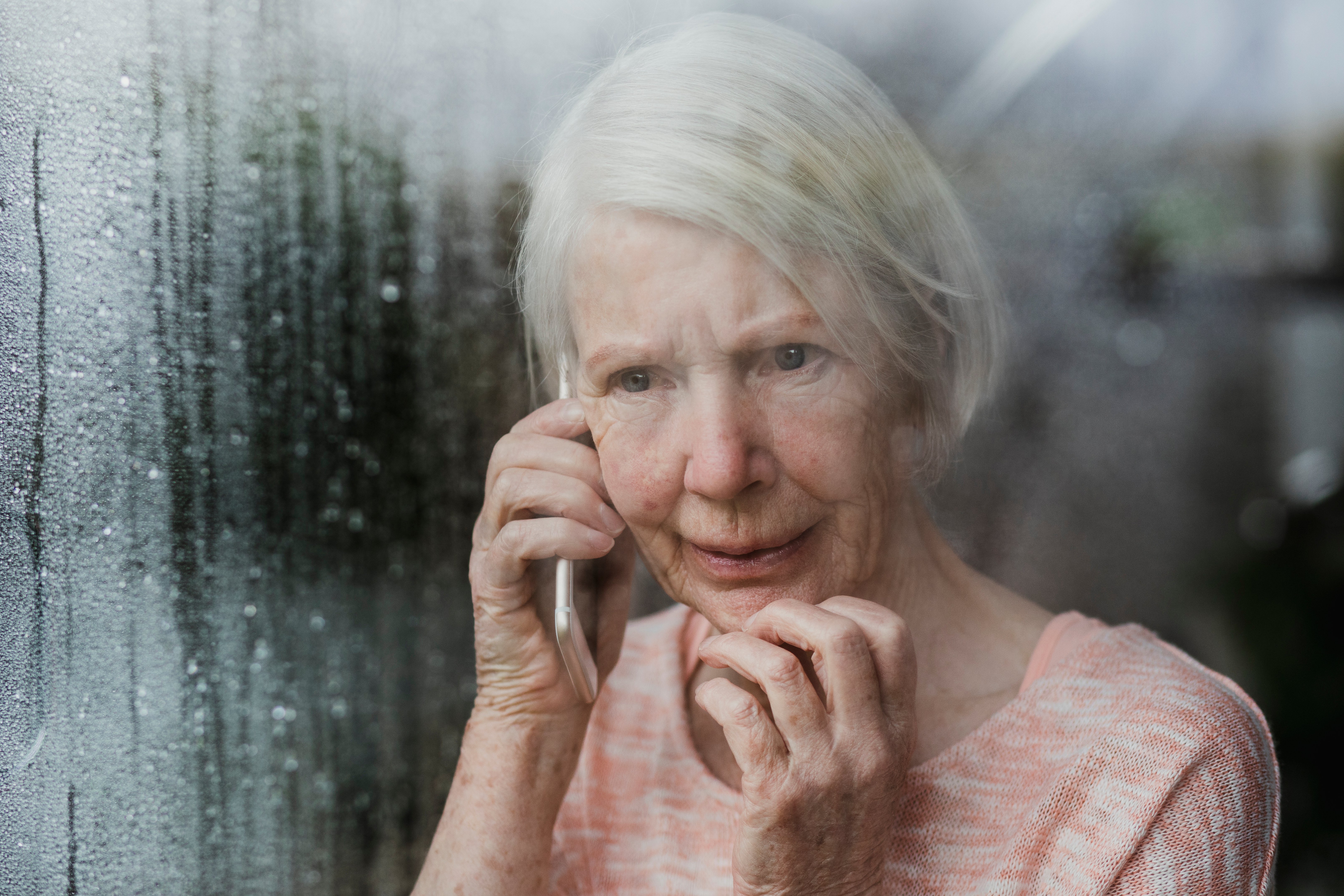 An older woman, looking out the window whilst on the phone and condensation coming down the window 