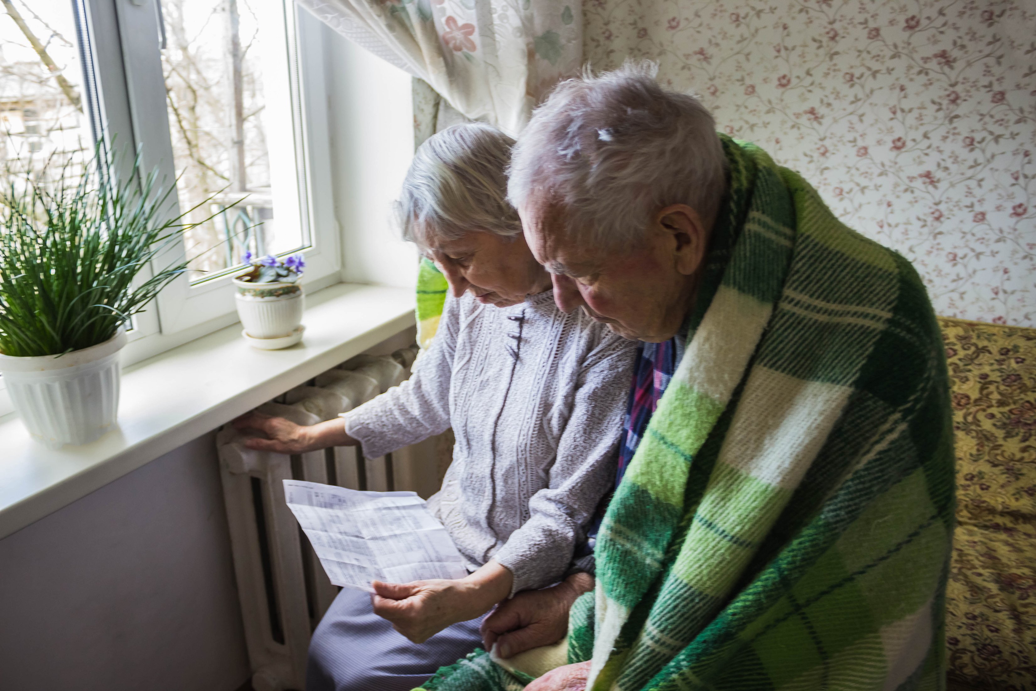 A older couple, the woman has her hand on the radiator and a energy bill, the husband is looking at the bill with a blanket around him